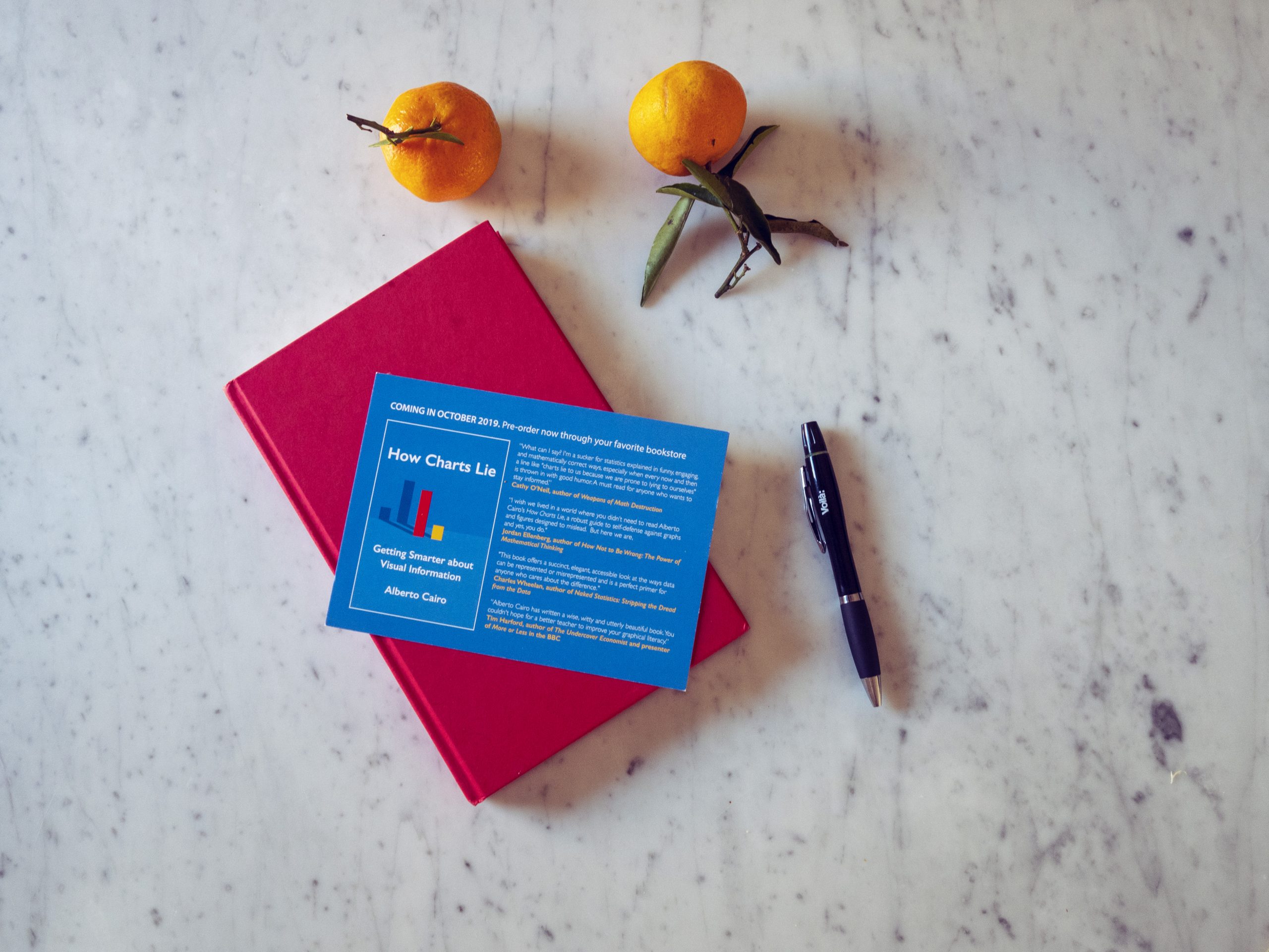 The book, closed, with a postcards on top introducing the book. Two clementines and a pen on the table next to it.