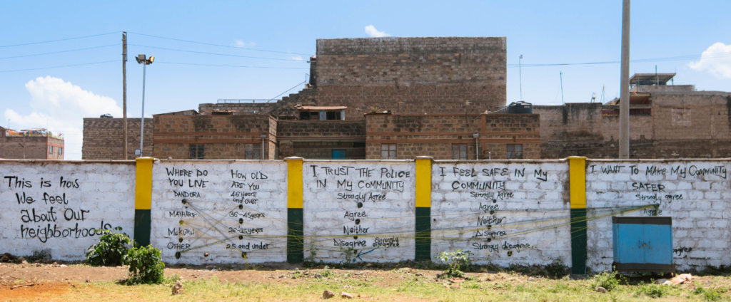 Un mur de ciment de 1,80 m de haut devant des bâtiments en briques dans un quartier informel du Kenya. Le mur est peint en blanc, est séparé par quatre colonnes vertes et jaunes, et porte une visualisation de données sous la forme de coordonnées parallèles avec des câbles traversant le mur tout en étant attachés à l'une des options de chaque catégorie. Le sujet est peint sur la gauche : «Voici ce que nous pensons de nos quartiers». Les catégories sont les suivantes : 1) Où habitez-vous, 2) Quel âge avez-vous, 3) Je fais confiance à la police de ma communauté, 4) Je me sens en sécurité dans ma communauté, 5) Je veux rendre ma communauté plus sûre. A six feet high cement wall in front of brick buildings in an informal settlement in Kenya. The wall is painted white, is separated by four green and yellow columns, and has a data visualization on it in the form of parallel coordinates with cables going across the wall while being attached to one of the options in each category. The topic is painted on the left: This is how we feel about our neighborhoods”. The categories are 1) Where do you live, 2) How old are you, 3) I trust the police in my community, 4) I feel safe in my community, 5) I want to make my community safer.