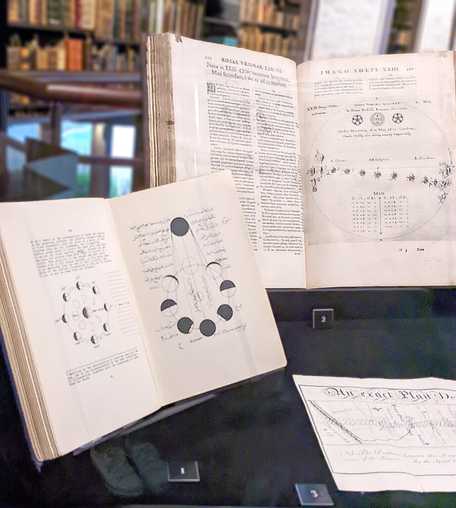 Open books arranged in a glass cabinet. One on the left has diagrams of moon phases, a set of circles arranged in a circle on the page, with various degrees of shading in black. Opposite the circles is another circle completely in black, representing the sun. Circles closest to the sun are least shaded, and the circle opposite the sun is completely shaded. Straight lines connect the sun and each shaded portion at different angles.