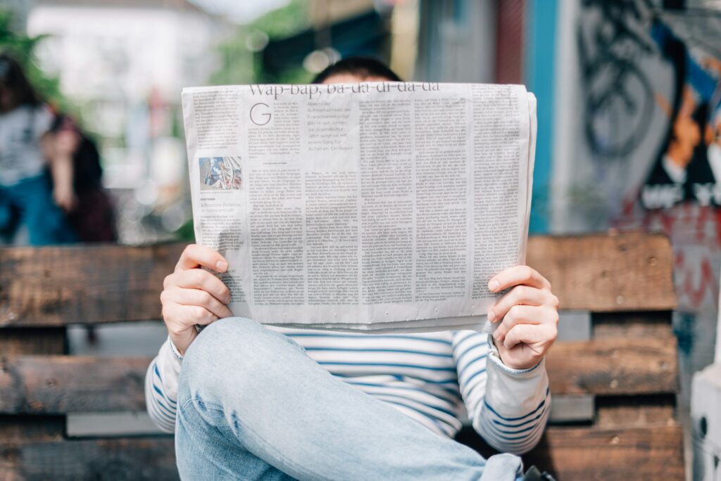 A man sitting on a wooden park bench, hiding behind the newspaper he's reading.