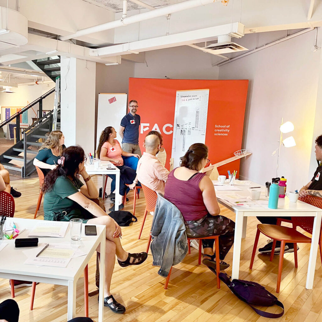 Francis Gagnon giving a training session to half a dozen people. Everyone is seated two by two at tables, and Francis listens to them while standing in front of a whiteboard with a drawing of a chart.