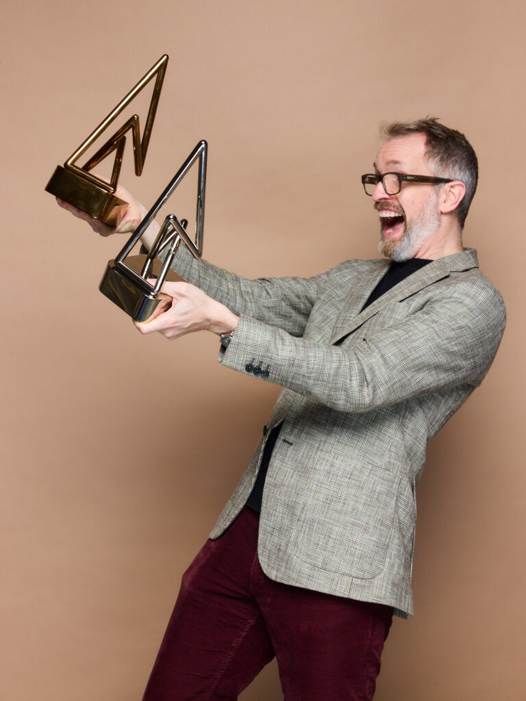 Francis poses on a monochrome background with one silver trophy in one hand and a bronze one in the other, in front of the event photographer. Turning to his right, as if he is talking to someone, he raises the prizes in the air with euphoric joy on his face. The trophies take the form of large, geometric A's made of metal tubing mounted on pedestals.