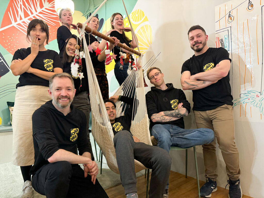 The entire team poses for the camera. In the foreground, Francis is kneeling, Julian is in the office hammock. To his left, Timour is sitting on a chair and Mathieu is standing. Behind the hammock, Jessie looks surprised, Celia and Marie-Blanche pause proudly, Patricia smiles and Estelle laughs. All are wearing black t-shirts with “Voilà” written on them.