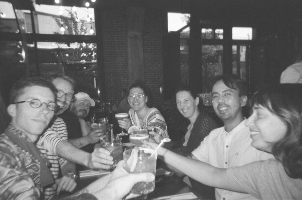 A black and white film picture of the team, gathered around a long table at a pizzeria. Everyone is smiling and raising their glasses in celebration after wrapping up major projects at Voilà:.