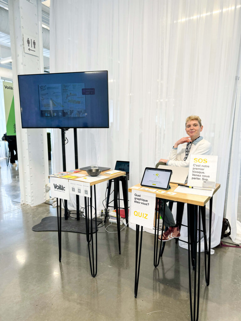 Une photo prise au Sommet climat Montréal. Celia est assise à notre kiosque et prend la pose. Notre kiosque est constitué de deux petites tables hautes sur lesquelles sont posés nos outils de promotion: des cartes, des flyers, un ipad, des stylos, etc. À droite de Celia, une grande télé est suspendue en hauteur et fait défiler une présentation de nos projets.