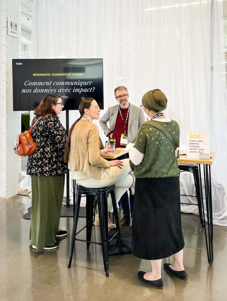 A photo taken at the Montreal Climate Summit. Francis organized a Braindate, an open conversation on the theme “How can we communicate our data with impact?”. Three women are standing in front of him, at our booth, in the middle of an exchange. The name of the theme is displayed on a large TV hanging to Francis' right.